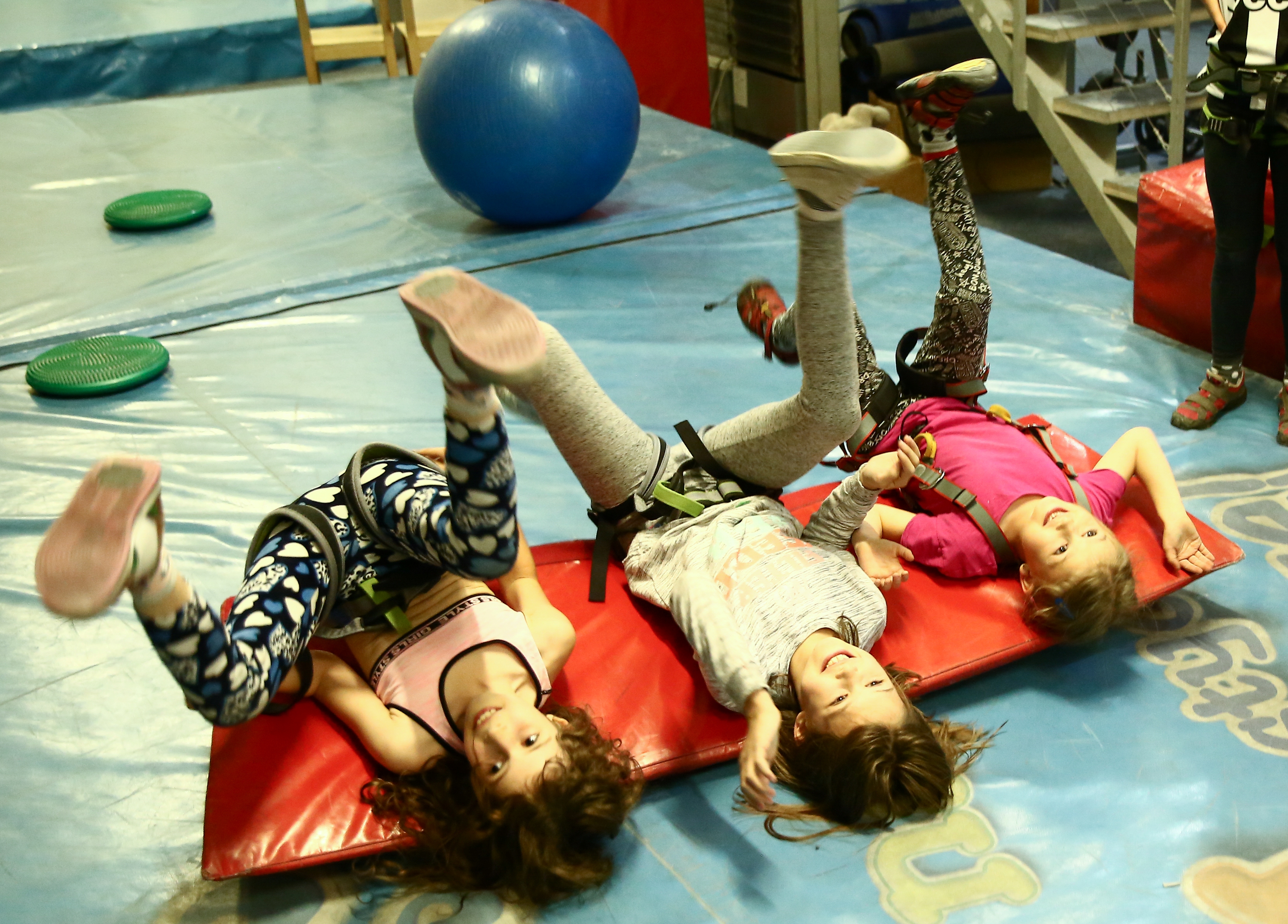 Trzy dziewczynki leżą na macie gimnastycznej i próbują zrobić fikołki
